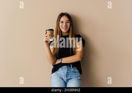 Lächelnde junge Frau trug Pullover stehend über beigen Hintergrund isoliert, die Tasse Kaffee zum Mitnehmen Stockfoto