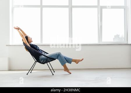 Junge hübsche Frau zu Hause sitzt auf modernen Stuhl vor dem Fenster entspannen in ihrem Wohnzimmer Stockfoto