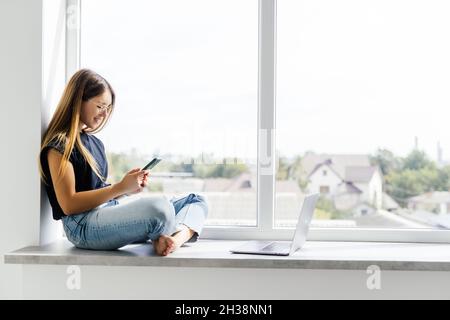 Glückliche Frau hält das Telefon, während sie zu Hause auf dem Fensterbrett sitzt. Stockfoto
