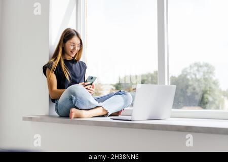 Glückliche Frau hält das Telefon, während sie zu Hause auf dem Fensterbrett sitzt. Stockfoto
