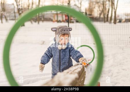 Kleinkind in grauem pelzigen Hut und blauem Einteiler, der mit Schnee auf p spielt Stockfoto