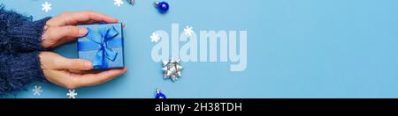 Kugeln und Schneeflocken sowie ein Geschenk für das neue Jahr in den Händen der Frau in dunklem Pullover auf blauem Hintergrund. Winter und Neujahr weihnachten Hintergrund. Stockfoto