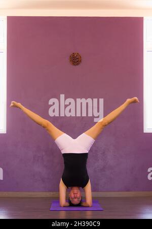 Junge Frau auf Kopfstand Pose mit gespaltenen Beinen. Weibliche Yogi in Sirsasana auf purpurfarbenem Wandstudio. Gleichgewicht, Gleichgewicht, Flexibilität, Bewegung, Training Stockfoto