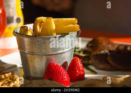 Kleiner Eimer aus silbernem Stahl mit Pommes frites mit Erdbeeren, Pilzen und Nüssen auf weißem Teller Stockfoto