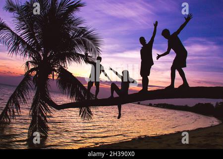 Silhouette einer Gruppe von Freunden, die in Balance auf einer liegenden Palme am Strand stehen, bei Sonnenuntergang auf der Insel Koh Phangan Stockfoto