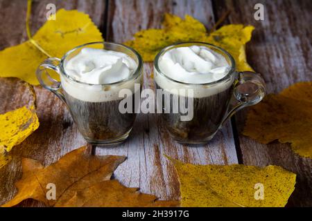 Zwei Tassen aus Glas mit Kaffeesahne auf dem Holztisch, der mit gelben Herbstblättern verziert ist Stockfoto