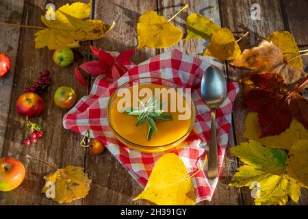 Kürbissuppe in einer Glasschüssel auf einem Holztisch, geschmückt mit bunten Herbstblättern und kleinen wilden Äpfeln Stockfoto