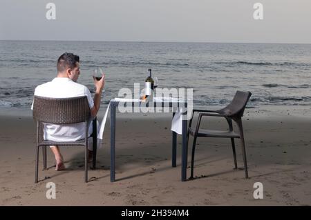Ein junger Mann in weißer Kleidung feiert ein romantisches Abendessen am Strand. Stockfoto