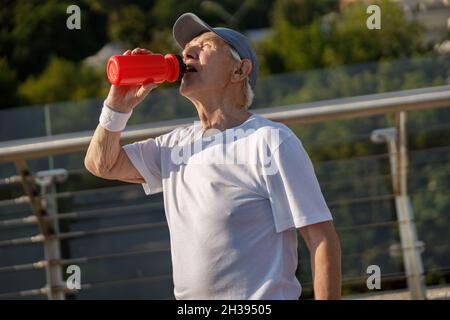 Älterer Herr trinkt Wasser aus der Flasche, die beim Training auf der Brücke steht Stockfoto