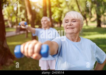 Starke ältere Frau macht Übungen mit Hanteltraining mit Ehemann im grünen Park Stockfoto