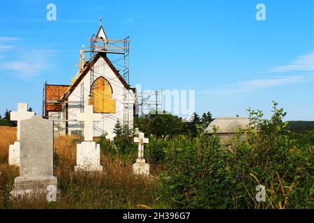 Eine alte Kirche, die repariert wird, mit Gerüsten, die um sie herum auf einem alten, überwucherten Friedhof errichtet wurden. Stockfoto