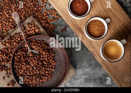 Drei Tassen mit Kaffee auf einem Holzbrett auf schwarzem Hintergrund. Eine mit Kaffeebohnen, die zweite mit gemahlenem Kaffee, die dritte mit schwarzem Kaffee Stockfoto