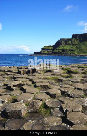 Blick auf schroffe grüne Klippen über blaues Wasser mit natürlichen vulkanischen Felsformationen im Vordergrund am Giant's Causeway in Nordirland. Stockfoto