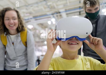 Positiver Junge mit VR-Brille, der während der COVID-Pandemie in der Nähe von Frau und Mann in einer Schutzmaske den Cyberspace erlebt Stockfoto