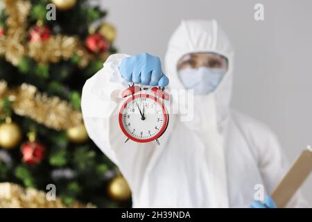 Arzt in schützendem Anti-Pest-Anzug mit Wecker in der Nähe des weihnachtsbaums Stockfoto