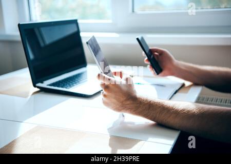 Die Hände eines Mannes halten zwei Telefone und einen Laptop auf dem Tisch. Moderne Technologie. Übertragung von Informationen von einem Gerät auf ein anderes Stockfoto