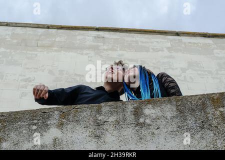 Ein fröhlicher Mann und eine fröhliche Frau unterhalten sich gegen eine graue Steinmauer. Ein junges heterosexuelles Paar. Ein Weibchen mit blauen Dreadlocks. Ein Männchen mit Brille. Aufnahme Stockfoto