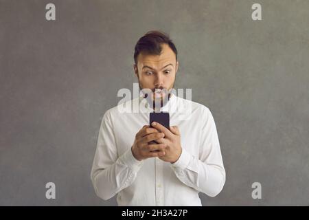 Der kaukasische Mann mit einem schockierten und peinlichen Ausdruck blickt auf ein Mobiltelefon in seinen Händen. Stockfoto