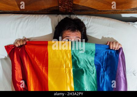 Ein junger Mann mittleren Alters, der im Bett liegt und sich mit der lgbtq-Flagge bedeckt, hat Angst Stockfoto