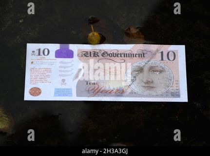 Eine gefälschte Banknote in einer Pfütze, als Darsteller von Ocean Rebellion als Premierminister Boris Johnson verkleidet und ein Oilhead das Segel eines kleinen Bootes anzünden, auf dem „die Zukunft Ihrer Kinder“ steht, während sie Stapel Geld am Ufer des Flusses Clyde in Glasgowm verbrennen In der Nähe des Standorts der bevorstehenden Cop26-Konferenz, die den Abbau des Ozeans beleuchtet. Die Szene stellt dar, was Rotester gesagt haben: „die britische Regierung hat keinen Sinn für die Bekämpfung des katastrophalen Klimawandels“. Bilddatum: Mittwoch, 27. Oktober 2021. Stockfoto