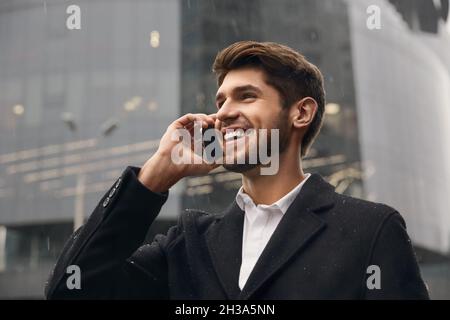 Junger lächelnder europäischer Geschäftsmann, der in der Stadt über ein Mobiltelefon spricht. Konzept der Fernarbeit. Moderner erfolgreicher Mann. Bärtiger, stilvoller Mann mit Mantel. R Stockfoto