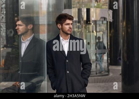 Junger seriöser europäischer Geschäftsmann, der neben einem Glasgebäude in der Stadt steht. Konzept des modernen erfolgreichen Menschen. Bärtig stilvollen Kerl mit Händen in den Taschen wea Stockfoto