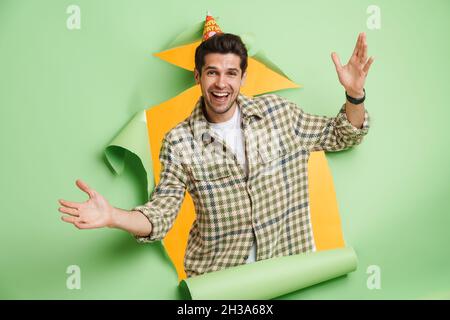 Lächelnd aufgeregt jungen beiläufigen weißen Mann Blick aus einem zerrissenen Papier Loch Hintergrund hält Arme ausgebreitet Geburtstagsfeier feiern Stockfoto