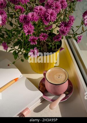 Kaffeetasse Schokolade Notizbuch schöne rosa Herbst Chrysantheme Blumen in gelben Topf auf Fenster. Gemütliches Zuhause Stockfoto