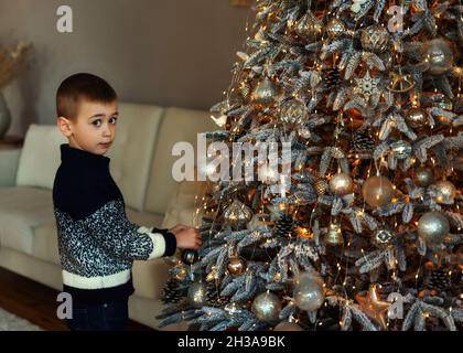 Ein Junge schmückt Weihnachtsbaum am Abend zu Hause. Ernsthaft arbeitsorientiert Kind Junge in einem Strickpullover Blick auf die Kamera. Familie mit Kindern feiern Wint Stockfoto