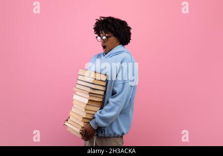 Überfordert schwarze Teenager-Student hält großen Stapel von Büchern, mit vielen Hausaufgaben auf rosa Hintergrund Stockfoto