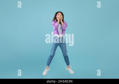 Schockierender Verkauf. Überrascht erstaunt junge asiatische Dame springen in der Luft und berühren Gesicht mit Erstaunen, blauer Hintergrund Stockfoto