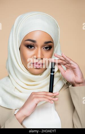Junge muslimische Frau in Hijab hält Mascara isoliert auf beige Stockfoto