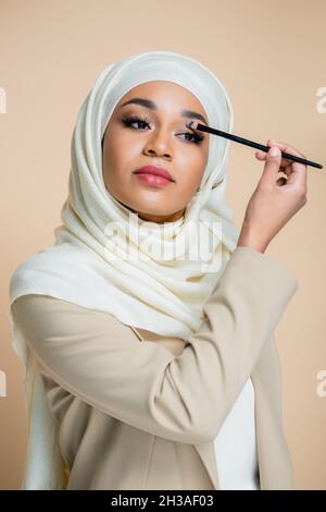 Junge muslimische Frau im Hijab, die einen auf Beige isolierten Lidschatten anwendet Stockfoto
