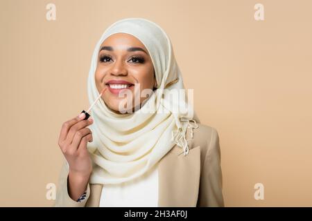 Glückliche muslimische Frau in Hijab Anwendung Lipgloss isoliert auf beige Stockfoto