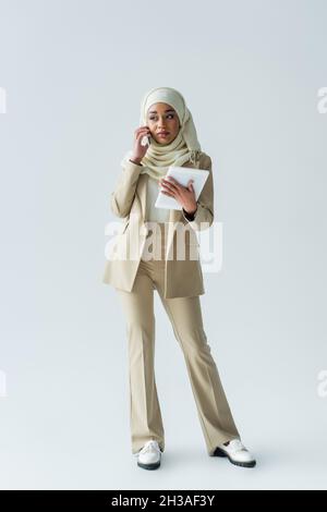 Die ganze Länge der jungen muslimischen Frau in Hijab hält digitale Tablette und spricht auf dem Smartphone auf grau Stockfoto