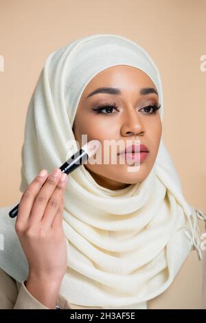 Junge muslimische Frau in Hijab, die Gesichtsfundament auf Beige isoliert anwendet Stockfoto