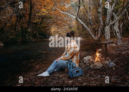 Ein Mädchen in einem karierten Hemd sitzt am Flussufer am Feuer auf einem Baumstamm. Stockfoto