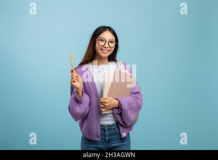 Ich kenne die Antwort. Aufgeregt asiatische Studentin in Brillen halten Notizblock und zeigen Bleistift nach oben, blauer Hintergrund Stockfoto