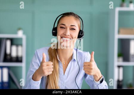 Fernschulung, Arbeit. Glückliche Dame im Headset zeigt Daumen nach oben, schaut und lächelt die Kamera im Büro an Stockfoto