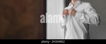 Ein Mann klammelt Knöpfe auf einem weißen Hemd mit seinen Händen im Licht vom Fenster. Stockfoto