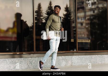 Afroamerikanische europäische College-Studentin mit Brille und Laptop-Computer geht ein junger Mann an einem sonnigen Tag, nea, getrost die Straße entlang Stockfoto