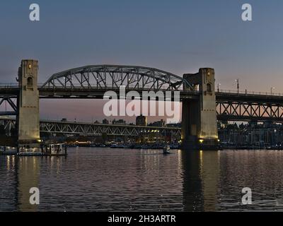 Vorderansicht der Burrard Street Bridge in der Innenstadt von Vancouver, die sich über die Bucht von False Creek erstreckt, mit beleuchteten Säulen im Abendlicht und rosa Himmel. Stockfoto