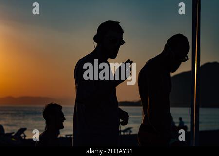 Silhouetten eines DJs und junger, cooler Männer, die am Strand bei Sonnenuntergang am Sevan-See, Armenien, feiern Stockfoto