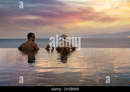 Romantischer Kurzurlaub - Pärchen trinken Champagner und genießen den wunderschönen Sonnenuntergang im Pool im tropischen Resort Stockfoto