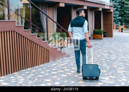 Der junge arabische Geschäftsmann Student kam mit dem Koffer in der Hand zum Restaurant, lächelte Kamera an, hält an und schaut weg. Stockfoto