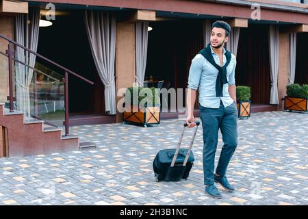 Der junge arabische Geschäftsmann Student kam mit dem Koffer in der Hand zum Restaurant, lächelte Kamera an, hält an und schaut weg. Stockfoto