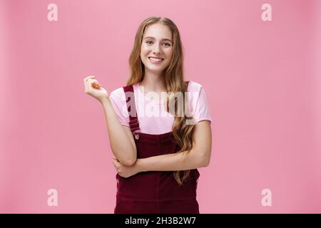 Nettes Mädchen in trendigen Overalls mit langen welligen Frisur und Licht Make-up stehen in lässiger Pose und lächeln fröhlich bei Kamera hören teilnehmen Stockfoto