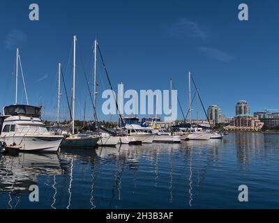 Blick auf den Yachthafen mit den Anlegebooten, die sich an sonnigen Tagen auf der glatten Wasseroberfläche von Victorias Binnenhafen mit Skyline im Hintergrund spiegeln. Stockfoto