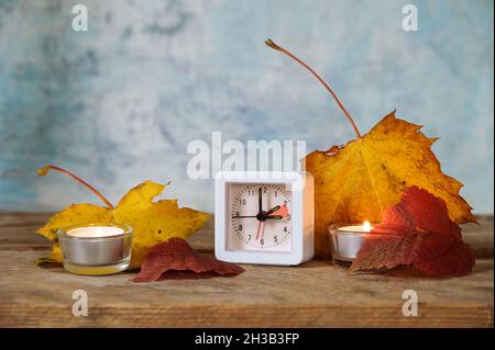 Wechsel von Sommer- auf Winterzeit im Oktober, kleiner Wecker eine Stunde zurückgestellt, Herbstblätter und Kerzen auf rustikalem Holz, hellblauer Hintergrund, Stockfoto