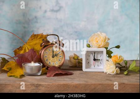 Uhrenwechsel, zwei Wecker, Retro-Stil und modern, zeigt Winter- und Sommerzeit, Herbstblätter und Blumen auf rustikalem Holz, hellblauer Rücken Stockfoto